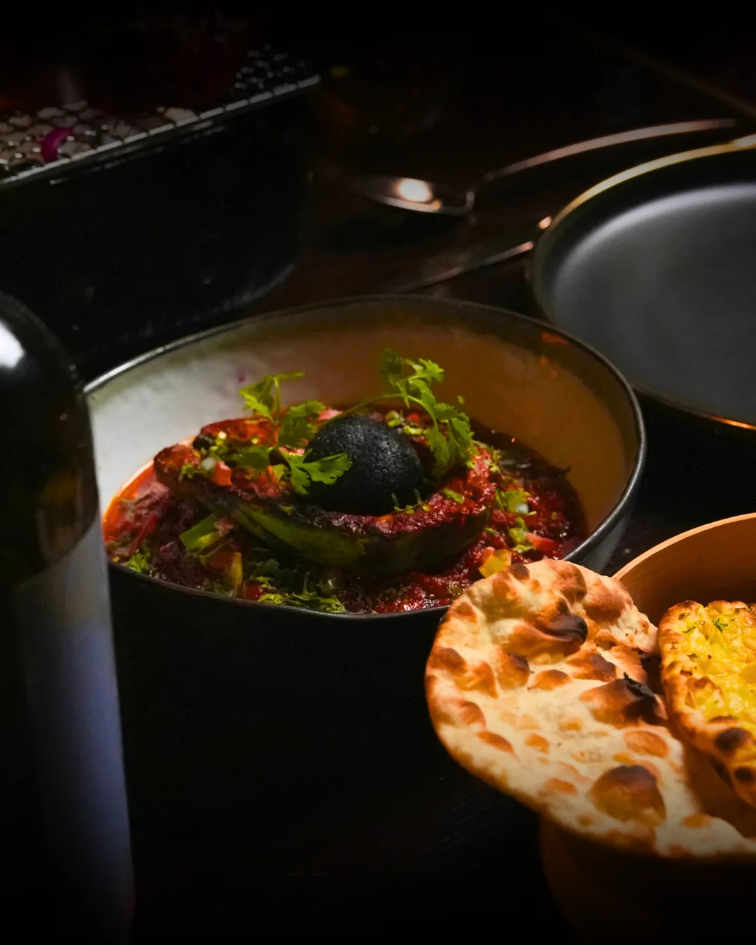 A curry of stuffed green chillies in a red masala, garnished with coriander, with naan on the side.
