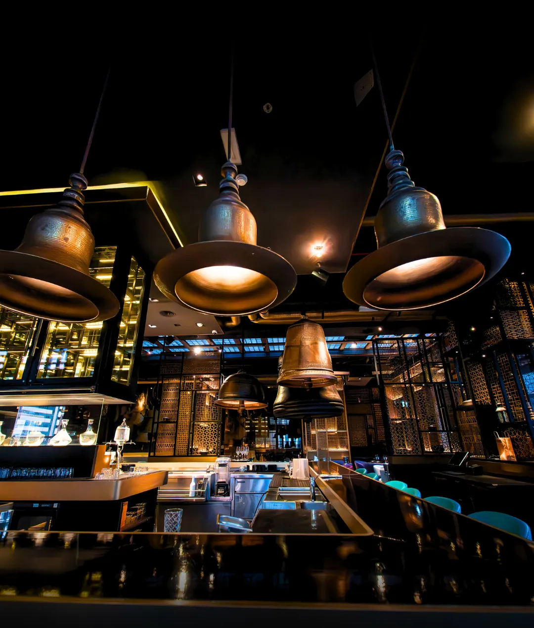 Three brass bell pendants hang low over the bar counter, gold lattice screens behind.
