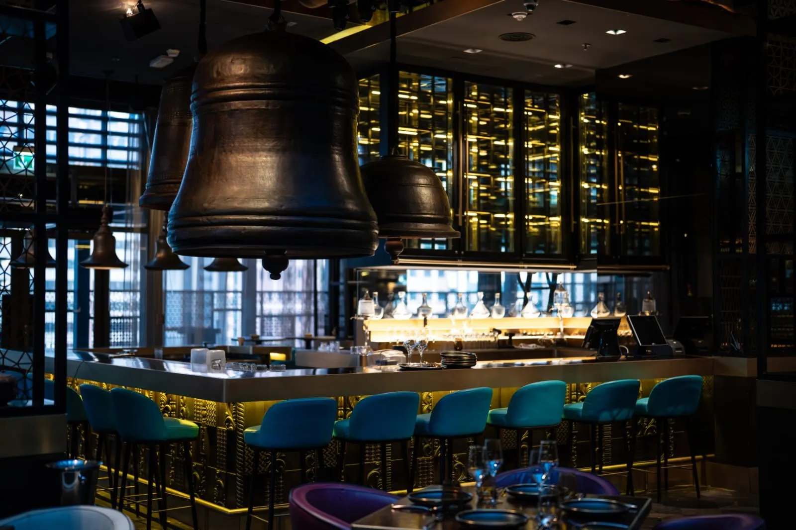 The backlit bar with a row of teal stools, a wall of bottles and bell pendants above.