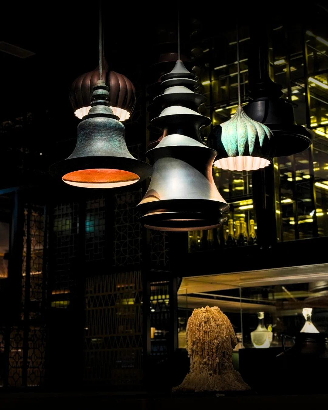 Sculpted pendant lamps in brass, verdigris and black glow above the bar, with bottles lit behind.
