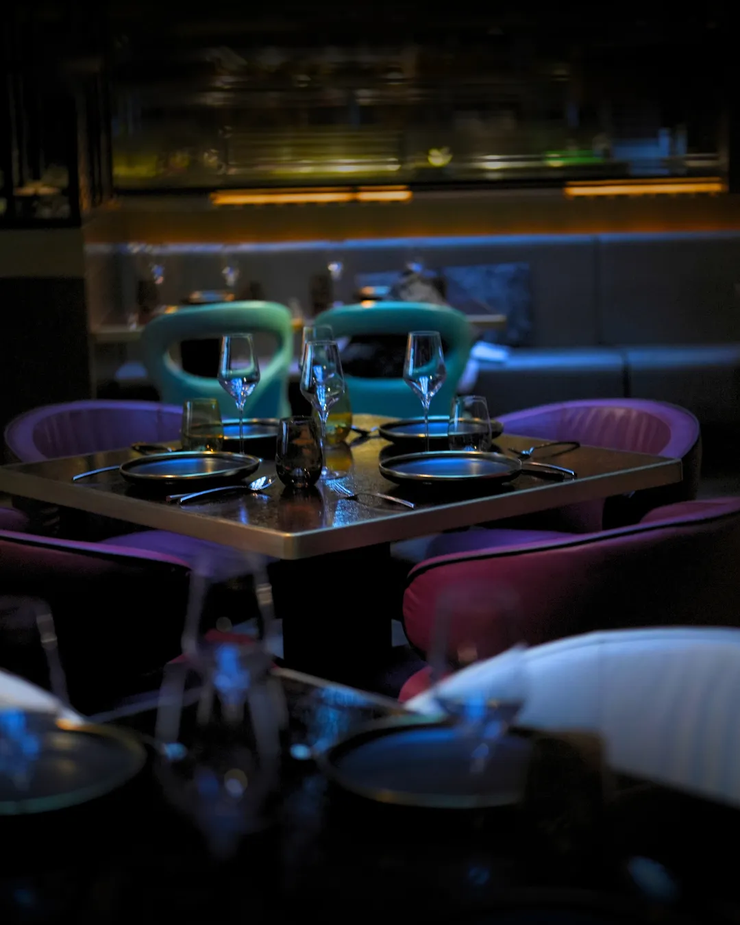 A table for four laid with black plates and crystal glasses between plum and teal chairs.